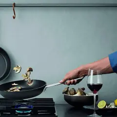 En hand steker svamp i en kolstålspanna på en gasspis. Bredvid står ett glas rödvin och en skål med svamp.