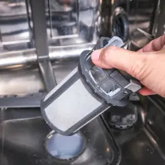 En hand tar ut ett smutsigt filter ur en diskmaskin. Ja, man kan diska rostfritt stål i diskmaskin, men filtret behöver rengöras.