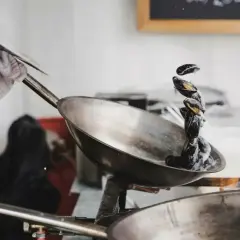 En hand med en trasa håller i ett handtag på en rostfri stekpanna där blåmusslor steks i en krämig sås.