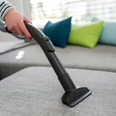 En hand dammsuger en grå soffa med en smal munstycke. Färgglada kuddar syns i bakgrunden.