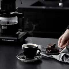 En hand sträcker sig efter chokladbitar bredvid en kopp rykande kaffe. Kaffebryggaren i bakgrunden ser ut att frysa kaffe för en perfekt start på dagen.