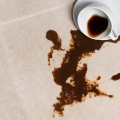 En vit kopp med kaffe har spillts ut på en ljus matta. Fläcken sprider sig olyckligt.