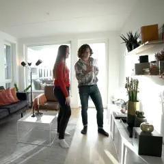 Två personer diskuterar inredning i ett modernt vardagsrum med en mörk soffa, vita möbler och hyllor med växter. Olika inredningsstilar syns i rummet.