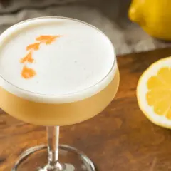 En läcker drink med skummig topp, dekorerad med en orange virvel. Bredvid ligger en citronskiva och en hel citron, redo att användas som sifon till drinkar.