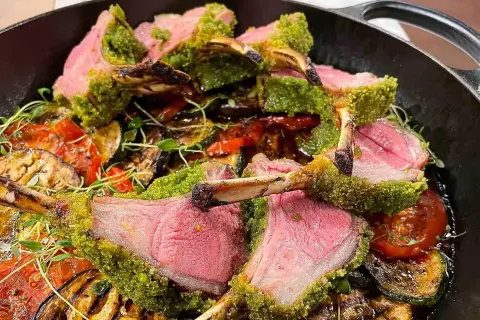 Saftigt lamm med perfekt lamm temperatur, täckt av gröna örter, serveras med grillade grönsaker i en gjutjärnspanna.