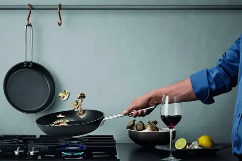 En hand steker svamp i en kolstålspanna på en gasspis. Bredvid står ett glas rödvin och en skål med svamp.