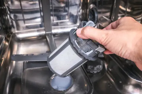 En hand tar ut ett smutsigt filter ur en diskmaskin. Ja, man kan diska rostfritt stål i diskmaskin, men filtret behöver rengöras.