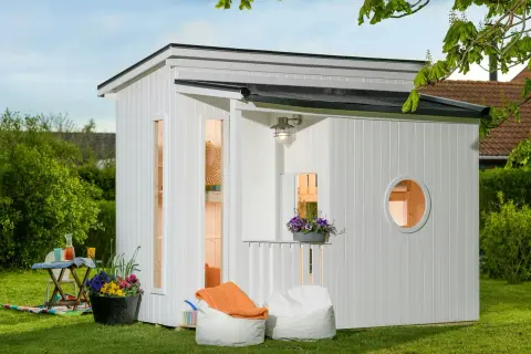 En vit lekstuga liten med fönster och en rund öppning. Uteplats med sittpuffar och blommor.