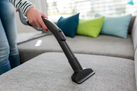 En hand dammsuger en grå soffa med en smal munstycke. Färgglada kuddar syns i bakgrunden.
