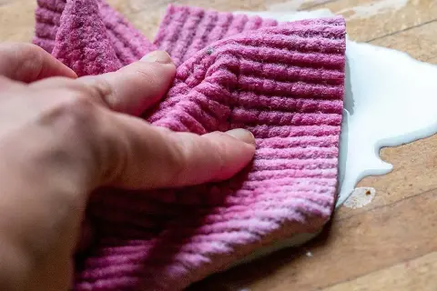 En hand håller en rosa trasa för att torka upp en vit vätska på en träyta.