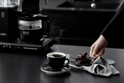En hand sträcker sig efter chokladbitar bredvid en kopp rykande kaffe. Kaffebryggaren i bakgrunden ser ut att frysa kaffe för en perfekt start på dagen.
