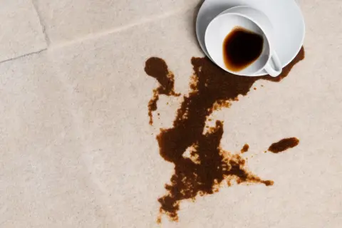 En vit kopp med kaffe har spillts ut på en ljus matta. Fläcken sprider sig olyckligt.