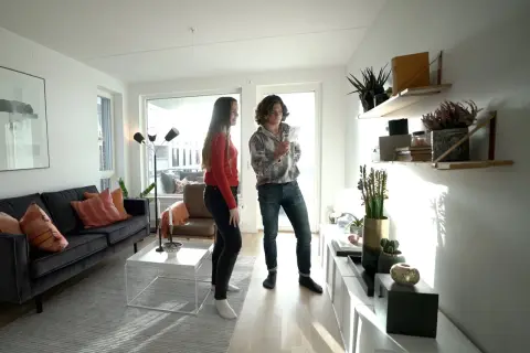 Två personer diskuterar inredning i ett modernt vardagsrum med en mörk soffa, vita möbler och hyllor med växter. Olika inredningsstilar syns i rummet.