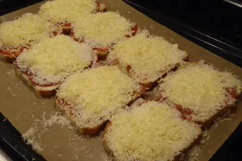 Flera smörgåsar med ost, tomat och skinka ligger på en plåt, redo för **toast i ugn**.