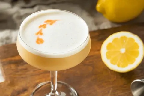 En läcker drink med skummig topp, dekorerad med en orange virvel. Bredvid ligger en citronskiva och en hel citron, redo att användas som sifon till drinkar.