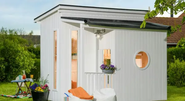 En vit lekstuga liten med fönster och en rund öppning. Uteplats med sittpuffar och blommor.