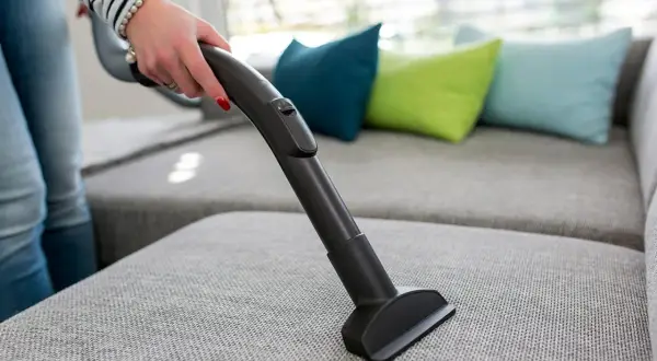 En hand dammsuger en grå soffa med en smal munstycke. Färgglada kuddar syns i bakgrunden.