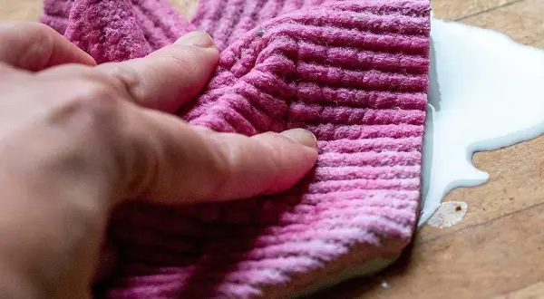 En hand håller en rosa trasa för att torka upp en vit vätska på en träyta.