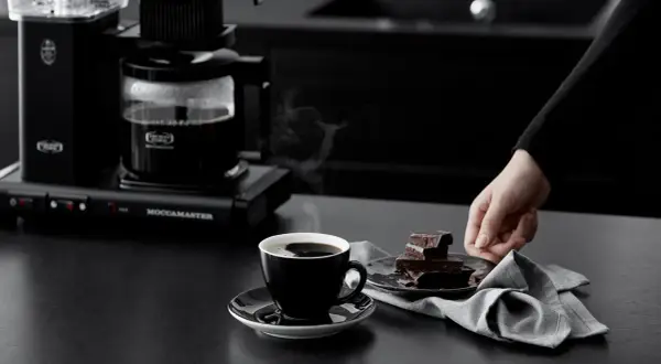 En hand sträcker sig efter chokladbitar bredvid en kopp rykande kaffe. Kaffebryggaren i bakgrunden ser ut att frysa kaffe för en perfekt start på dagen.