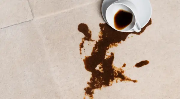En vit kopp med kaffe har spillts ut på en ljus matta. Fläcken sprider sig olyckligt.