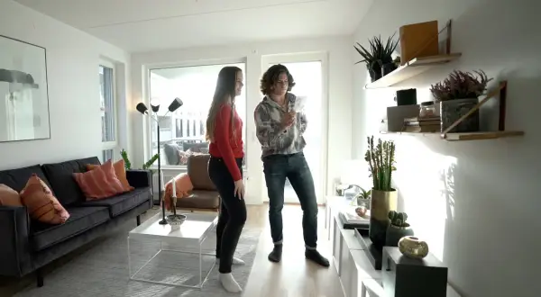 Två personer diskuterar inredning i ett modernt vardagsrum med en mörk soffa, vita möbler och hyllor med växter. Olika inredningsstilar syns i rummet.