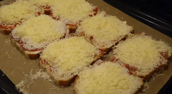 Flera smörgåsar med ost, tomat och skinka ligger på en plåt, redo för **toast i ugn**.