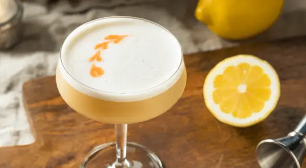 En läcker drink med skummig topp, dekorerad med en orange virvel. Bredvid ligger en citronskiva och en hel citron, redo att användas som sifon till drinkar.
