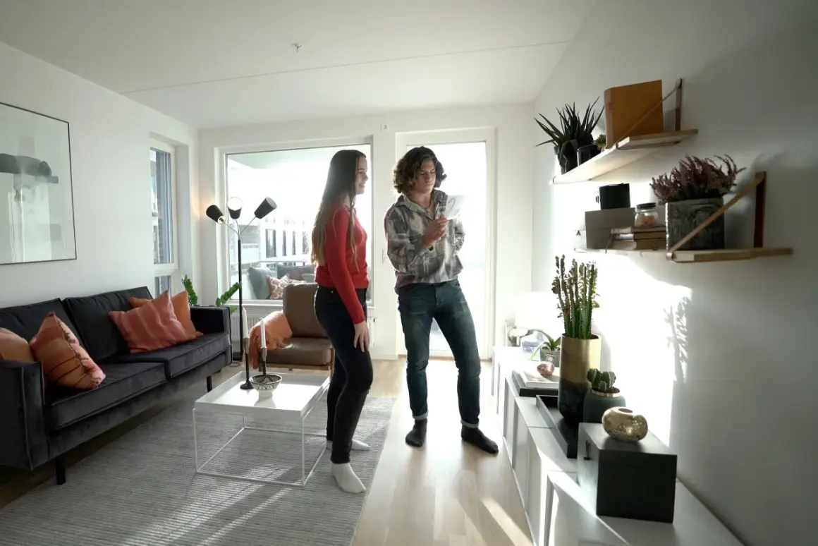 Två personer diskuterar inredning i ett modernt vardagsrum med en mörk soffa, vita möbler och hyllor med växter. Olika inredningsstilar syns i rummet.