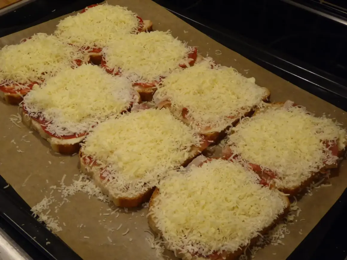 Flera smörgåsar med ost, tomat och skinka ligger på en plåt, redo för **toast i ugn**.