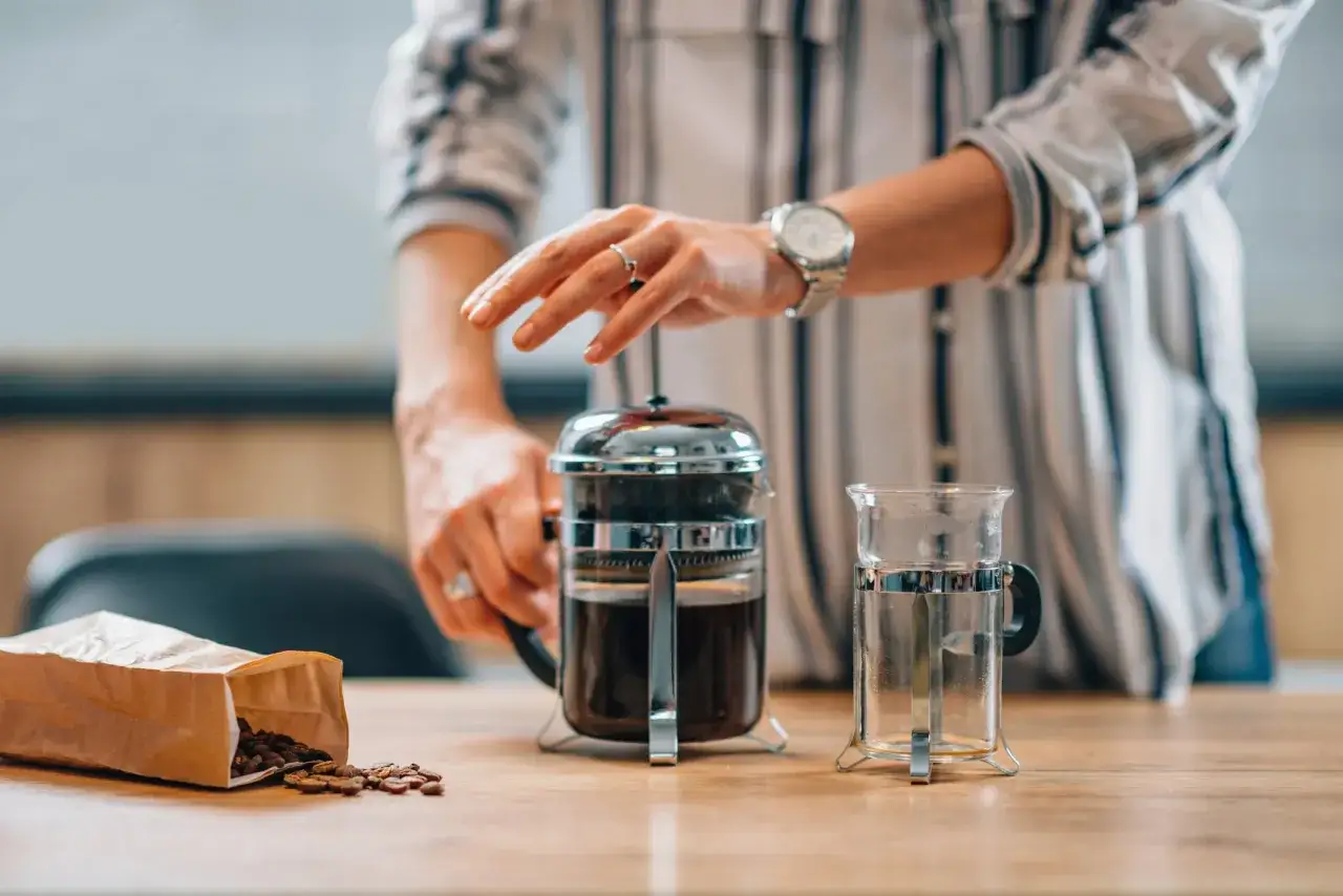 Personen förbereder sig för att brygga kaffe manuellt med en presskanna. Kaffebönor ligger bredvid.