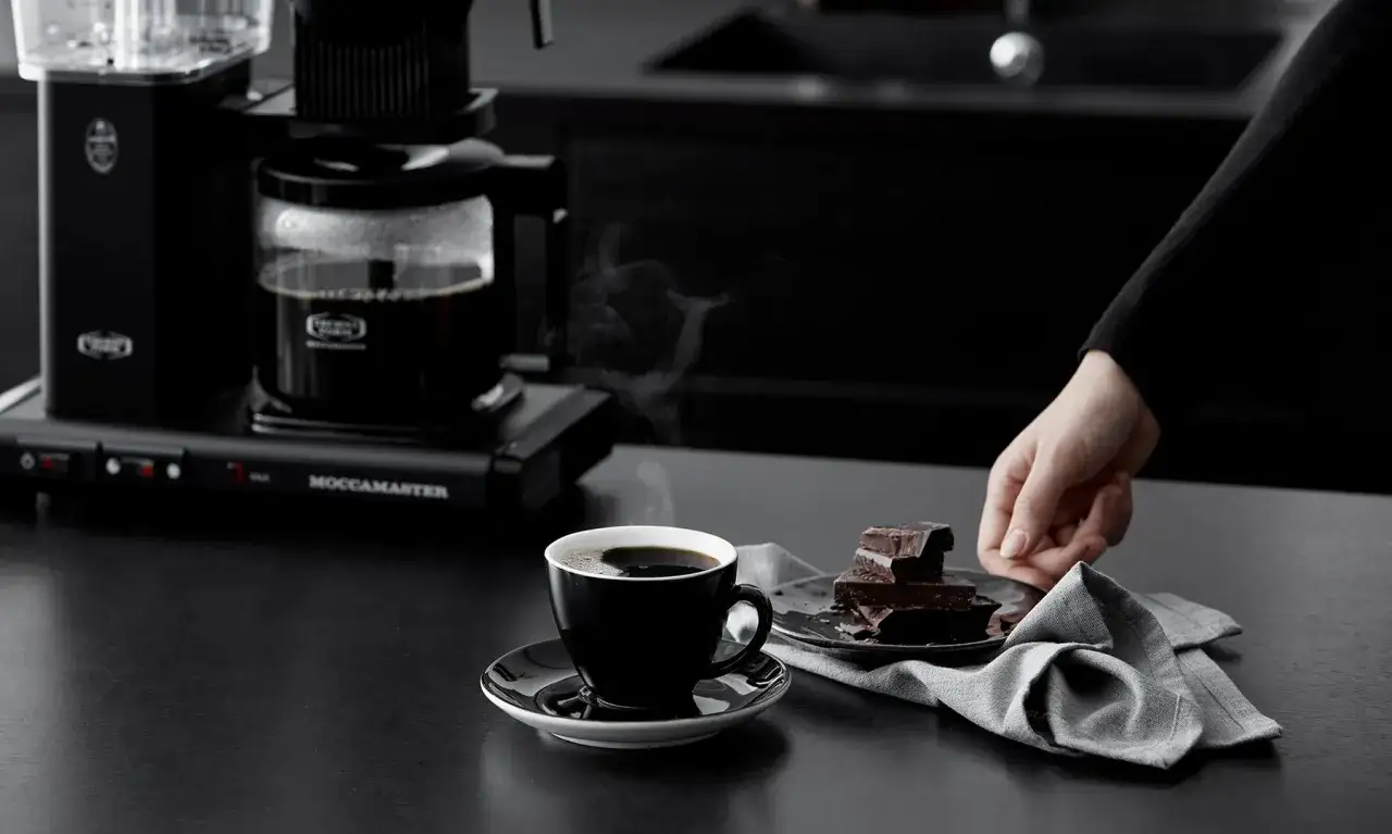 En kopp rykande kaffe och chokladbitar på en mörk bänk. Kaffebryggaren Moccamaster står i bakgrunden. Perfekt för att inte frysa kaffe.