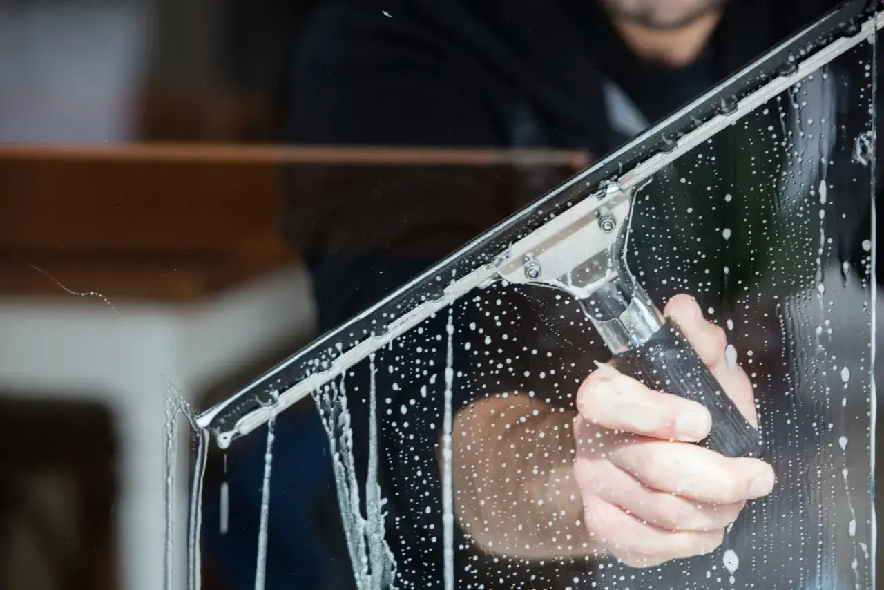 En hand håller en fönsterskrapa med såpbubblor som rinner nerför glaset. Resultatet blir bästa fönsterputs.