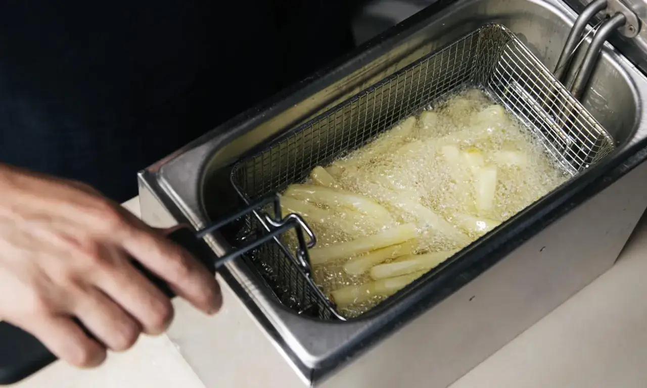 Pommes frites friteras i en kastrull med bubblande olja. En hand håller i korgen med pommes.