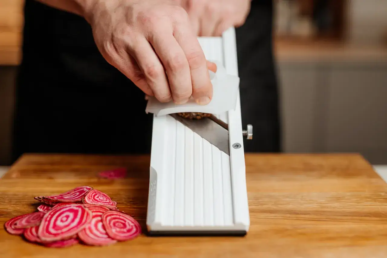 En hand använder en mandolin för att skiva rödbetor. Tunna, cirkulära skivor av mandolin grönsaker ligger på en träskärbräda.