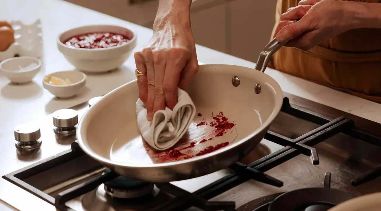En hand torkar ur en stekpanna med en trasa, rester av r&ouml;da b&auml;r syns. Ska man diska stekpannan med diskmedel?