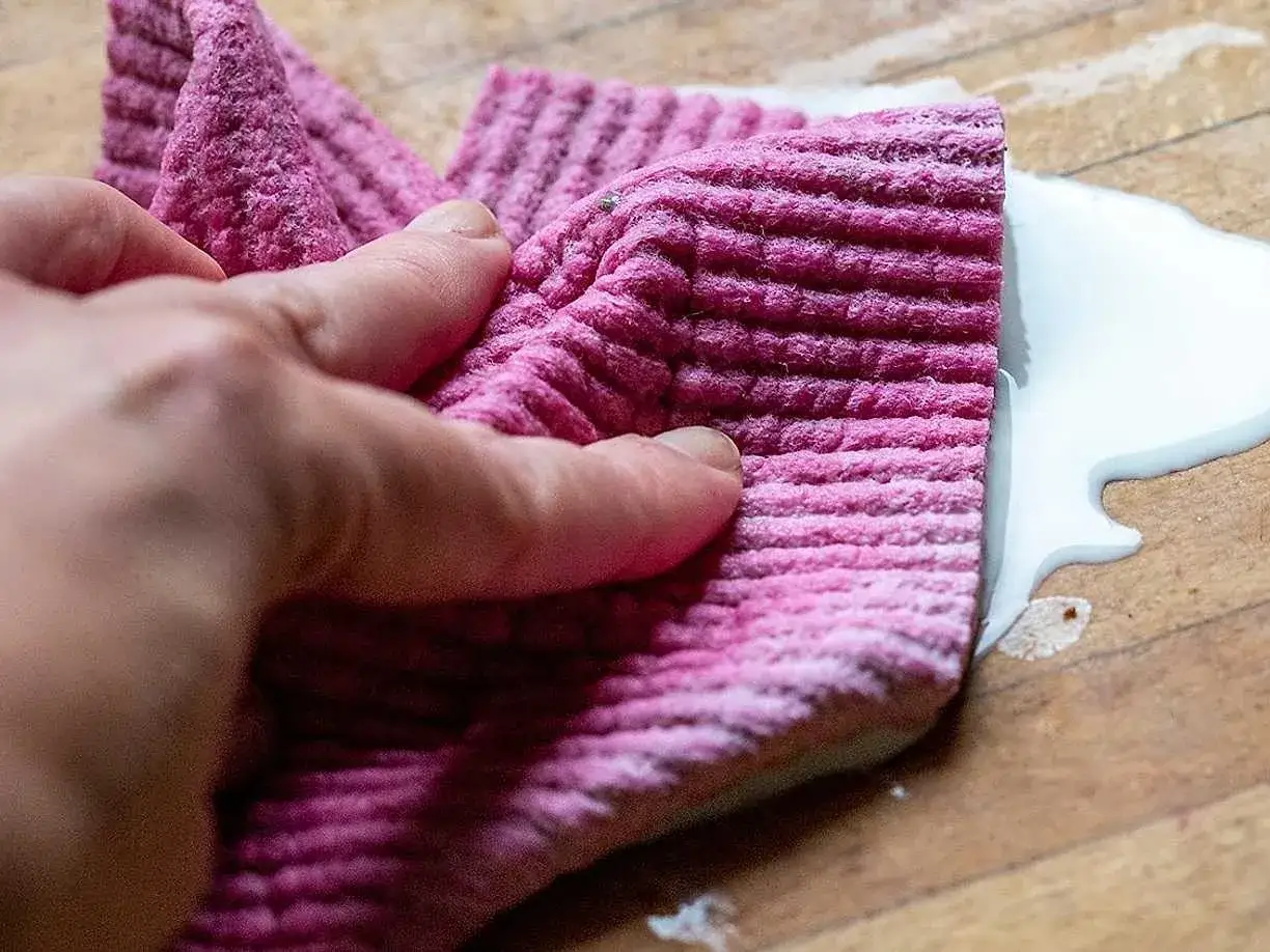 En hand håller en rosa trasa för att torka upp en vit vätska på en träyta.