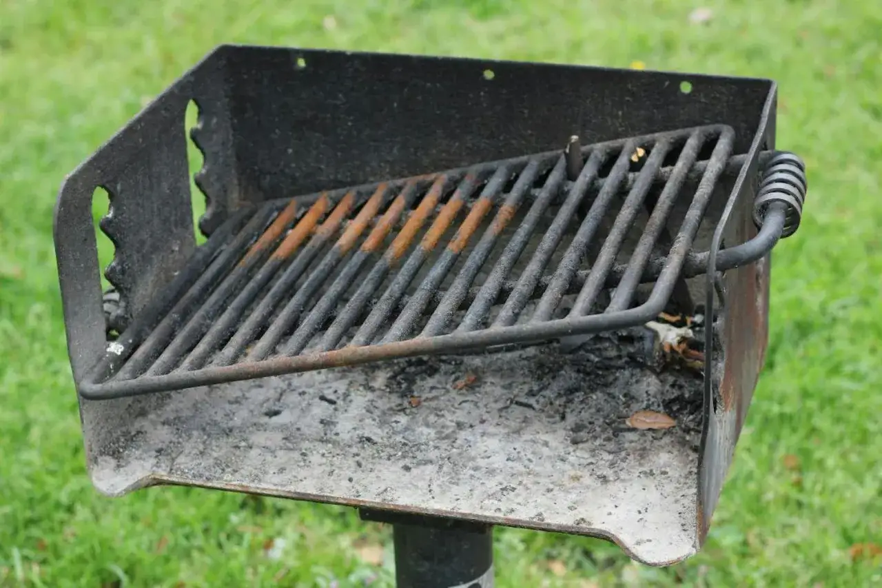 Ett rostigt grillgaller med rester av aska och sot. Grillen st&aring;r utomhus p&aring; en gr&auml;smatta.