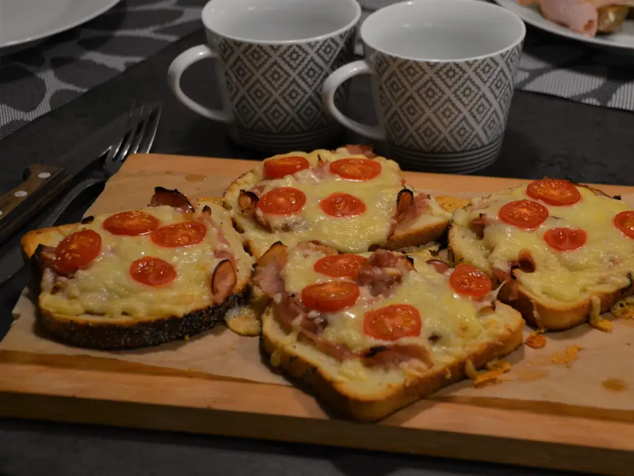 Fyra varma toast i ugn med ost, skinka och tomater ligger på en skärbräda. Två mönstrade muggar står bakom.