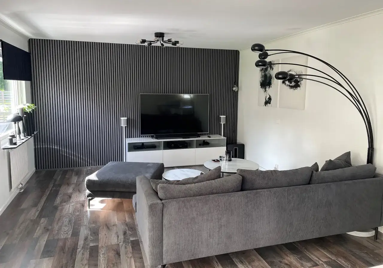 Modern inredning tv-rum med gr&aring; soffa, ribbad v&auml;gg och golv i tr&auml;imitation. En stor TV st&aring;r p&aring; ett vitt sk&aring;p.