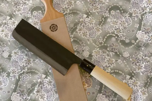 En vacker strigla kniv med mörkt blad och ljust trähandtag ligger på ett tyg med blomstermönster.