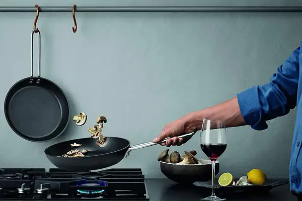 En hand steker svamp i en kolstålspanna på en gasspis. Bredvid står ett glas rödvin och en skål med svamp.