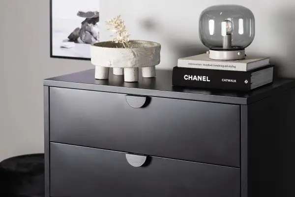 Svart byrå med två lådor, dekorerad med en kruka med torkad växt och böcker. Perfekt byrå sovrum inspiration.