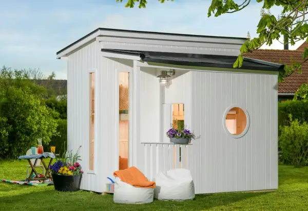 En vit lekstuga liten med fönster och en rund öppning. Uteplats med sittpuffar och blommor.