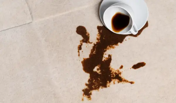 En vit kopp med kaffe har spillts ut på en ljus matta. Fläcken sprider sig olyckligt.
