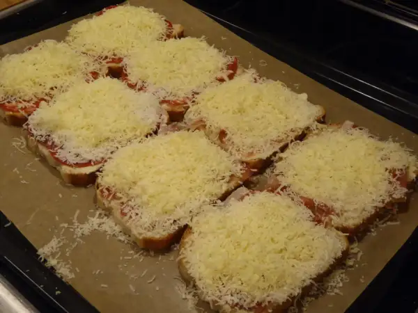 Flera smörgåsar med ost, tomat och skinka ligger på en plåt, redo för **toast i ugn**.